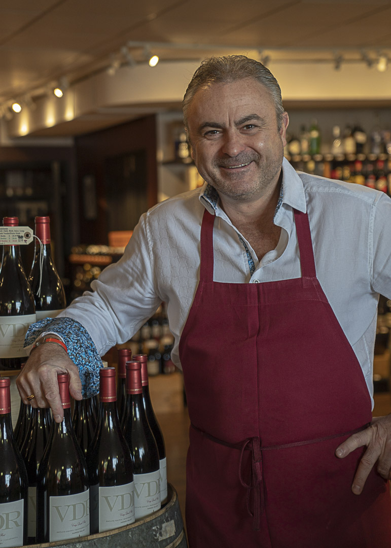 Indoor photo of Christophe Lhopitault, owner of Lake Side Emotions Wine Boutique, wearing a dark red apron, standing by a wooden barril on top of which bottles of red wine are presented to the customer.